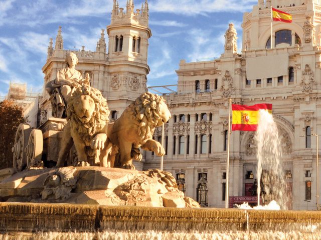 Statue of lions near a fountain, with Spanish flag and historic building in the background.